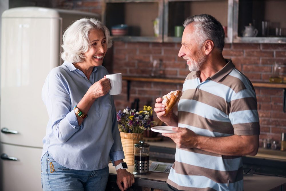 Happy elderly couple smiling at each other while drinking coffee In a good mood. Hunband tell jokes to his classy-looking wife in the kitchen